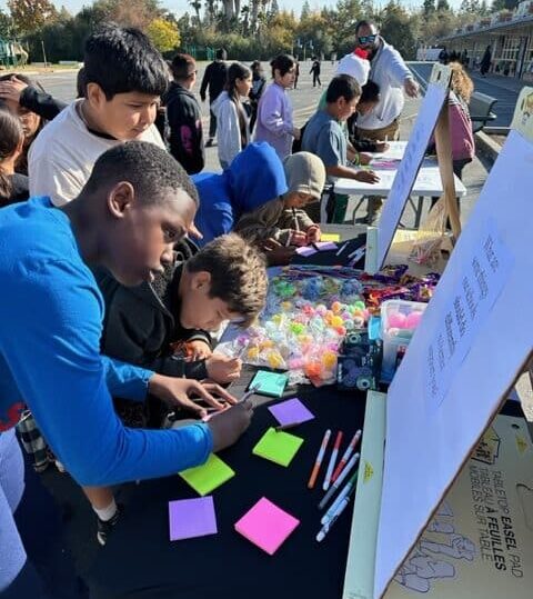 Elementary students gather around an outdoor table, writing ideas on colorful sticky notes and participating in a hands-on community engagement activity on a school campus.